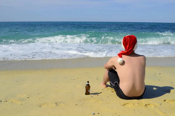 熱帯クリスマス サンタクロースの帽子の男が海岸でビールを飲む — ストック写真