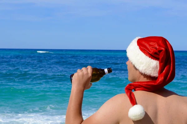 热带圣诞 一个戴着圣诞老人帽子的男人在海滨喝啤酒 — 图库照片