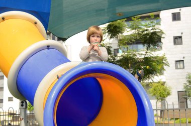 A little boy rides down a slide on a playground near the house. Yard in a modern neighborhood. The kid rejoices in summer and a sunny day. Concept: kindergarten, child care, behavior, entertainment, attractions