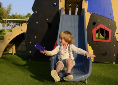 Küçük bir çocuk evin yakınındaki bir oyun parkında kaydırağın üzerinden kayıyor. Modern bir mahallede bahçe. Çocuk yazın ve güneşli bir günde sevinir. Konsept: anaokulu, çocuk bakımı, davranış, eğlence
