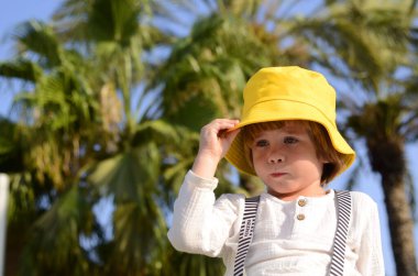 Portrait of a cute boy in a yellow panama hat under palms tree. A cute child rejoices in the summer. Concept: travel to the resort, holidays with children, kindergarten, holiday, birthday.