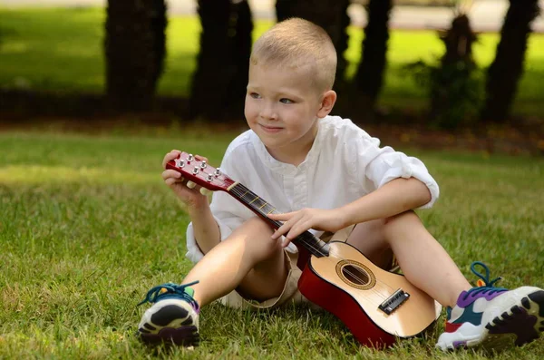 Little boy plays the acoustic guitar in the park. The child learns to play the uculele. Music lessons for children. Hawaiian guitar under a palm tree.