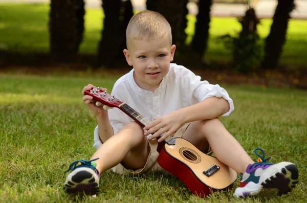 Little boy plays the acoustic guitar in the park. The child learns to play the uculele. Music lessons for children. Hawaiian guitar under a palm tree.