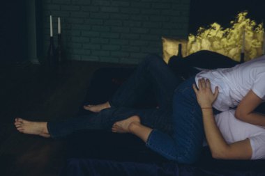 Beyaz tişörtlü ve kot pantolonlu çift karanlık bir odada, yatay bir yatakta yatağın üzerinde yatıyor.