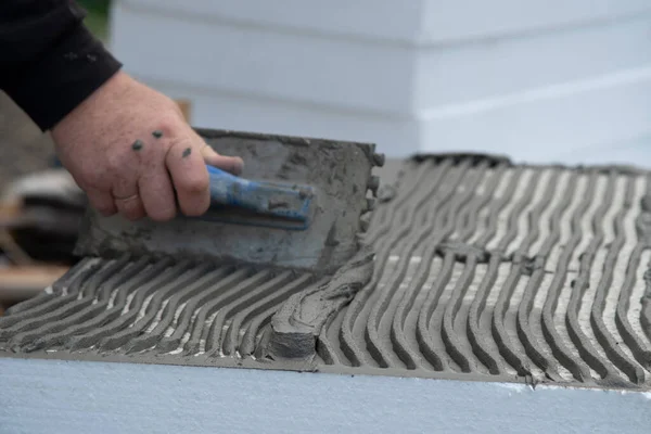 Close-up of worker hand, with trowel applying glue on white rigid polyurethane foam sheet for house insulation.