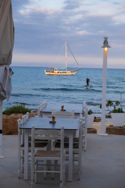 Seaside restaurant tables paddle surfer and a sailboat bathed in afternoon light in Potamos Liopetri fishing village in the Mediterranean island of Cyprus