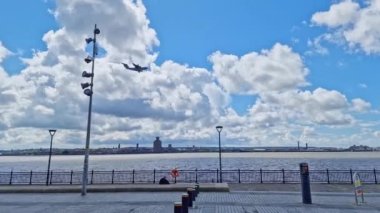 A military transport airplane flies by at the Liverpool waterfront of the Liverpool Docks as tourists stroll by on a spring afternoon