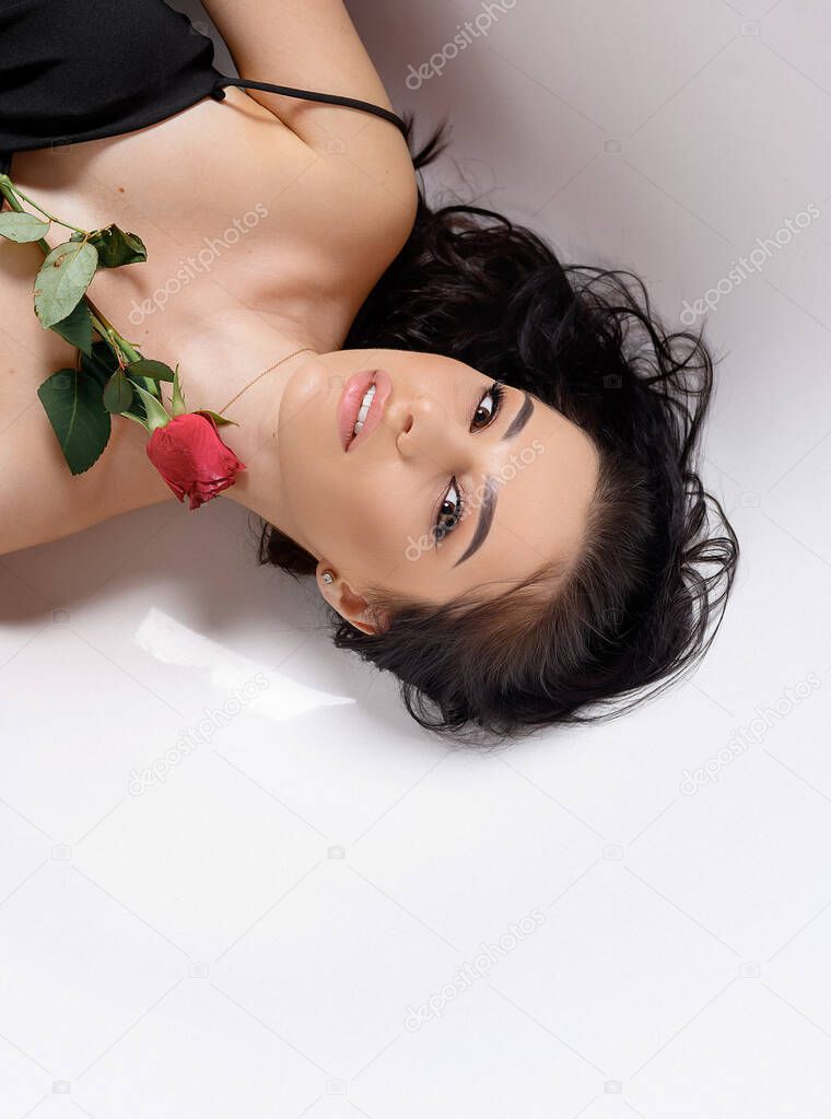 Young beautiful brunette girl in a black dress with a red rose lies in a white bathroom and looks into the frame