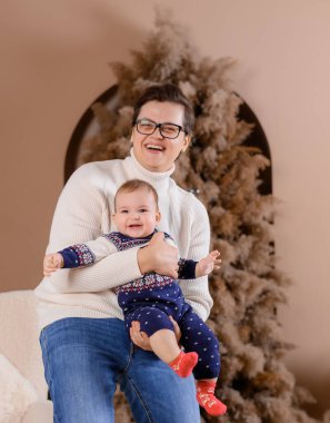 Genç bir aile babası ve oğlu yeni yıl ağacının arka planında şakalaşıp boş boş oturuyor.