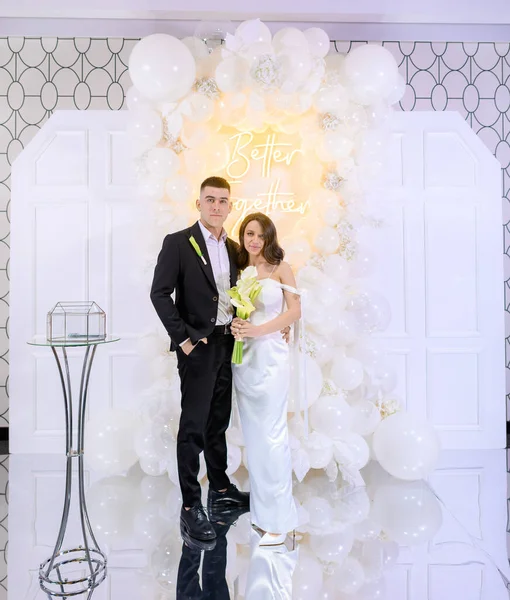 Beautiful newlywed couple posing on a background of photo decor with white balloons — Stok Foto