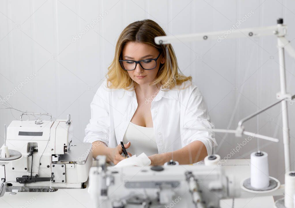 Young beautiful blonde hair seamstress cuts the fabric in the workplace.