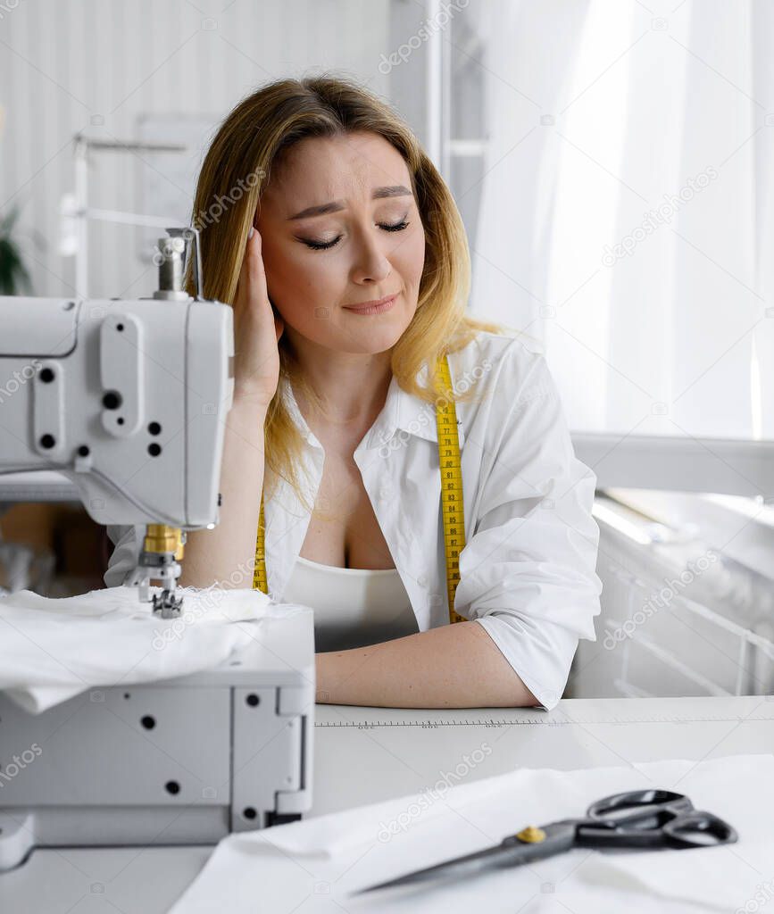 Young beautiful blonde hair seamstress sits at work and shows a headache