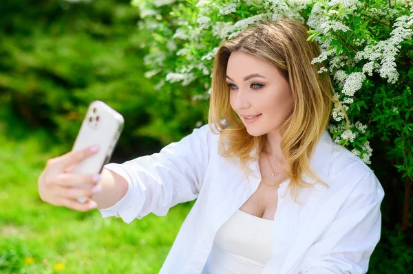 Beautiful blonde hair girl in a white shirt holds a phone and takes a selfie — Zdjęcie stockowe