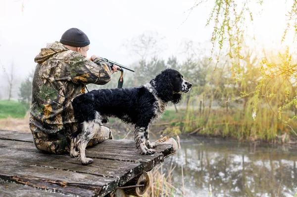 Männlicher Jäger mit russischem Spaniel sitzt auf einer Brücke und zielt mit einem Gewehr — Stockfoto