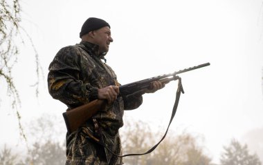 Omzunda silah olan yakışıklı bir adam sisli göle bakıyor.
