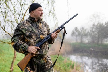 Omzunda silah olan yakışıklı bir adam sisli göle bakıyor.