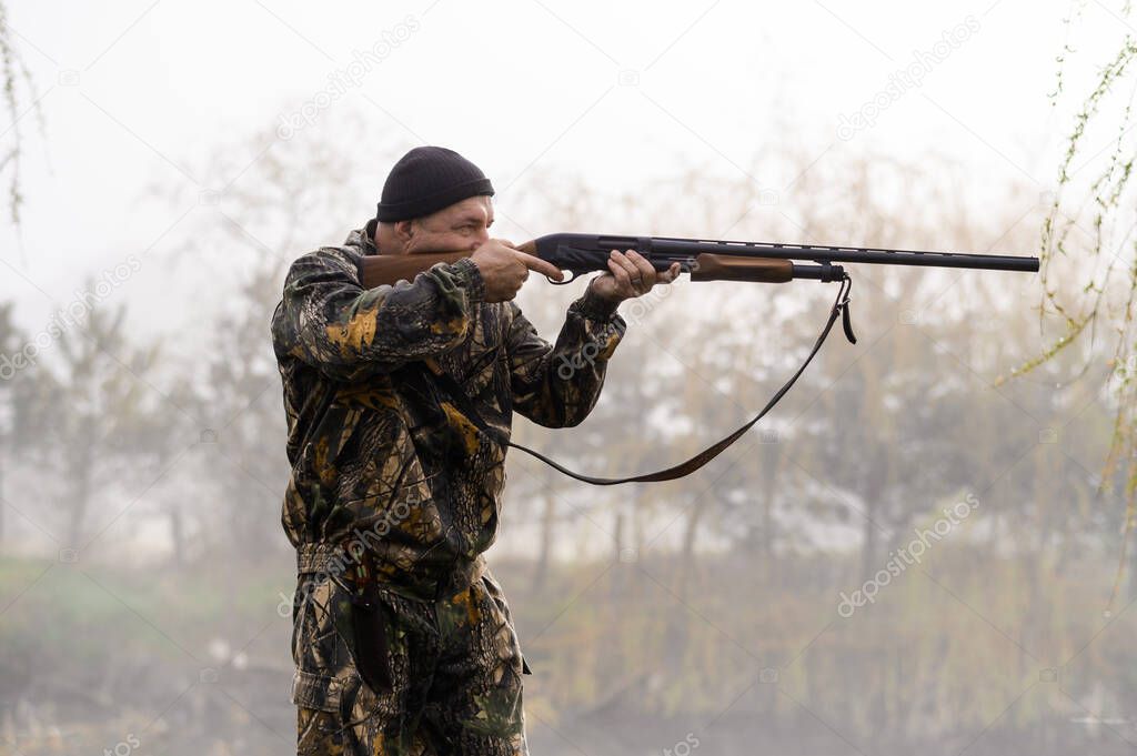 A male hunter with a gun hunts and aims against the background of reeds