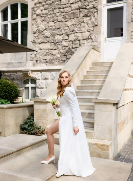 Beautiful blonde caucasian bride in a white dress with a slit posing — Stok fotoğraf