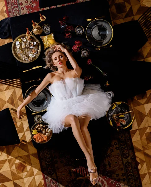 Beautiful caucasian blonde girl in a white dress lies on a set table — Stock Photo, Image