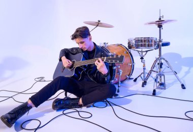 Genç adam akustik gitar çalıyor. Yerde oturuyor ve davul fıçısına yaslanıyor.