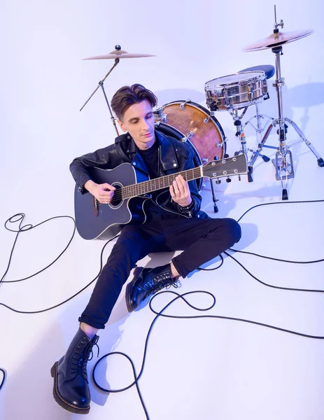 Young guy playing acoustic guitar sitting on the floor leaning on a drum barrel