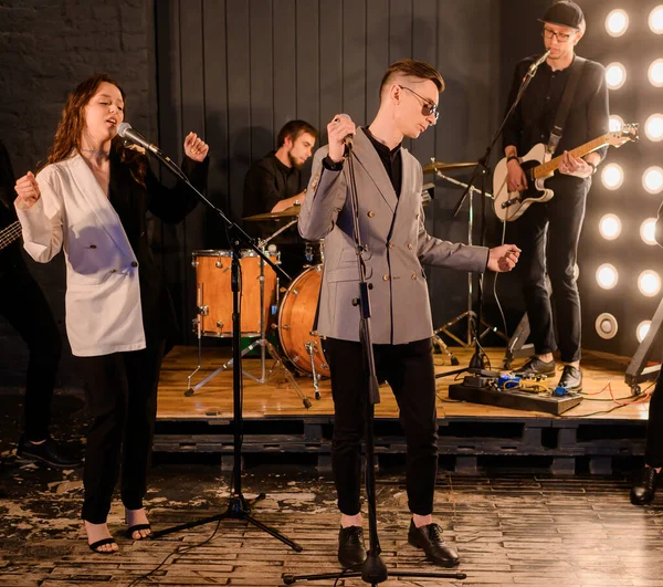 Two Caucasian vocalists singing against the backdrop of a band