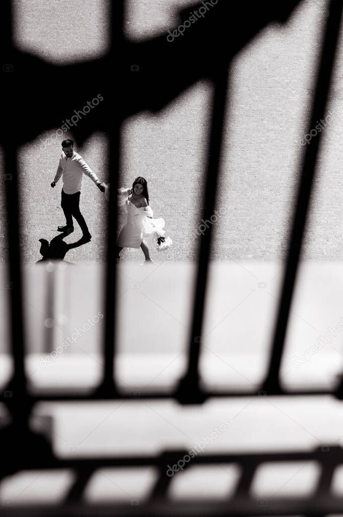 Beautiful young caucasian bride in white goes holding the hand of the groom. Shot through the window grille. Black and white photo