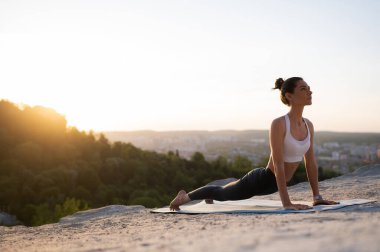 Güzel beyaz kız yoga yapıyor ve günbatımı ve şehir manzarasına karşı bir kayanın üzerinde tahta egzersizi yapıyor.