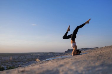 Güzel, genç, beyaz bir kız yoga yapıyor ve günbatımı ve şehir manzarasının arka planında bacaklarını bir kayaya çarpıştırarak amuda kalkıyor.