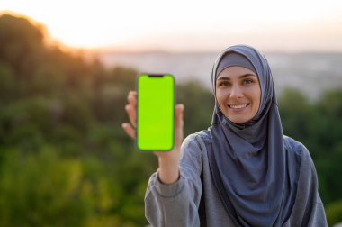 Güzel genç kız tesettürlü elinde yeşil ekranlı bir telefonla gün batımında manzaranın arka planında.