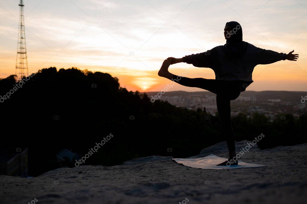 Silhouette of a beautiful young girl in a hijab meditates and practices yoga on a background of the landscape at sunset
