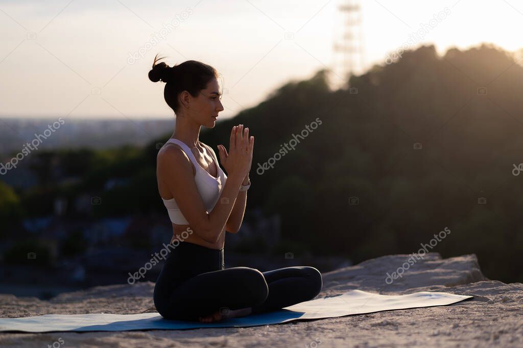 Beautiful young caucasian girl practicing yoga and meditating on a rock against the backdrop of sunset and cityscape