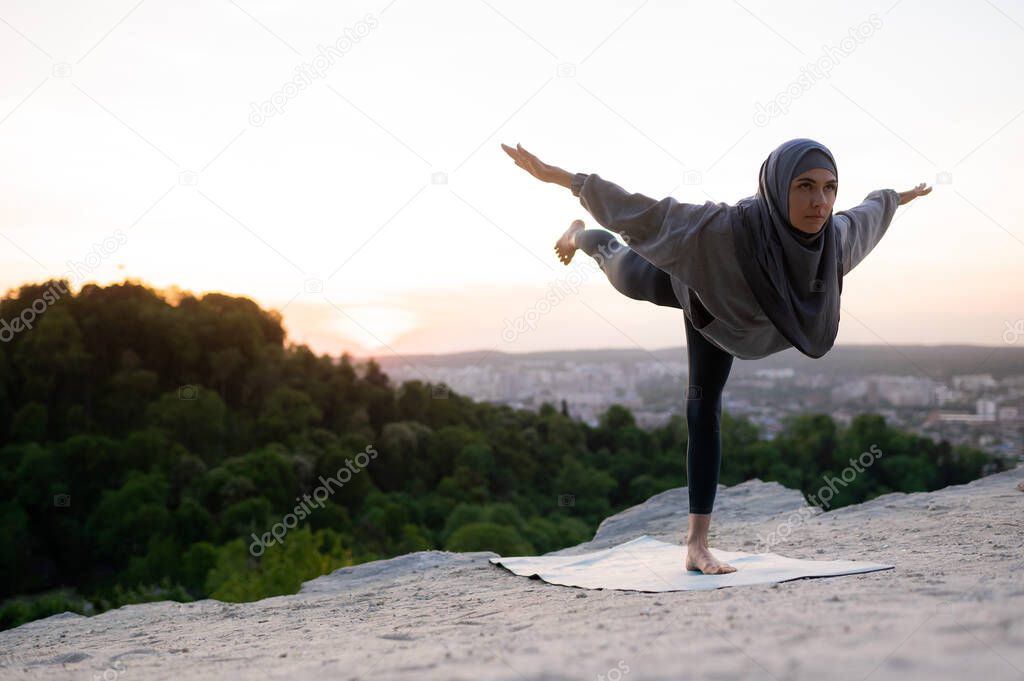 Beautiful young girl in hijab meditates and practices yoga on the background of the landscape at sunset