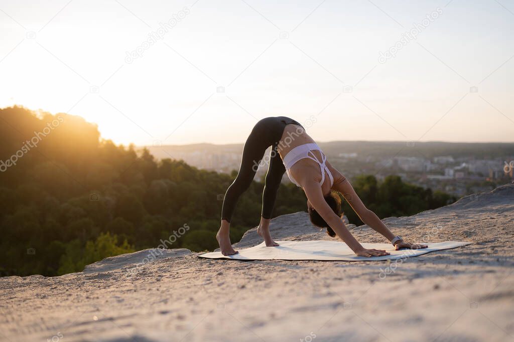 Beautiful young caucasian girl practicing yoga and performing asana on a rock against the backdrop of sunset and cityscape