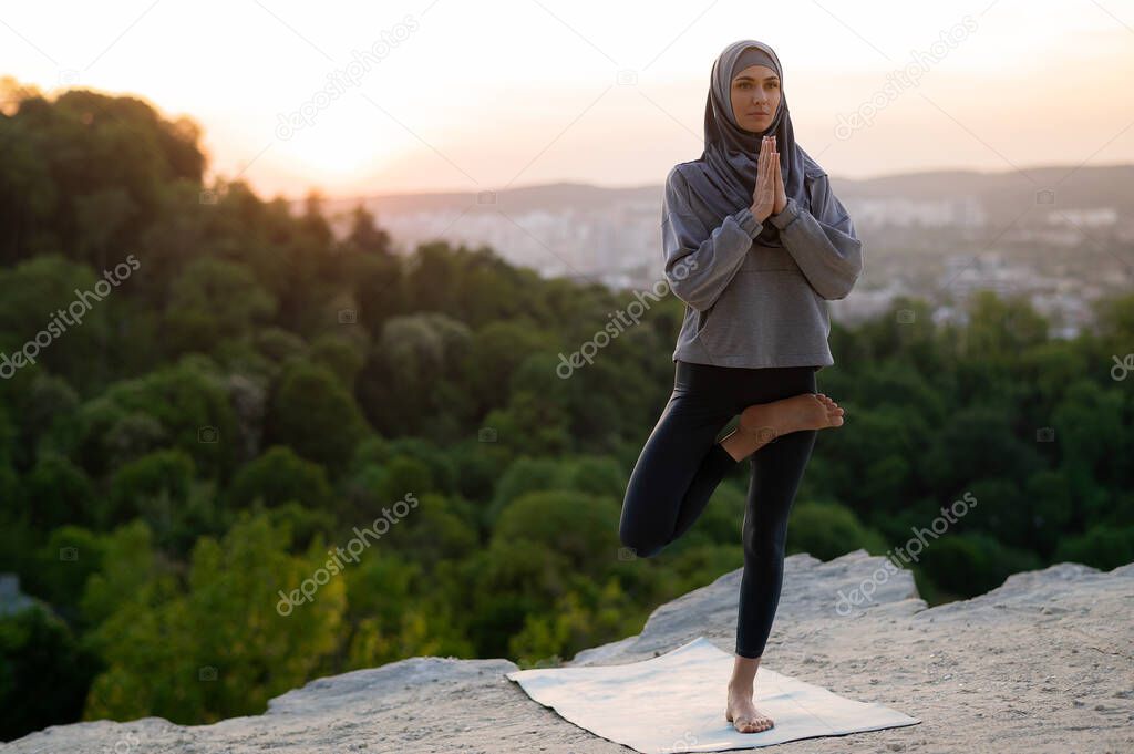 Beautiful young girl in hijab meditates and practices yoga on the background of the landscape at sunset