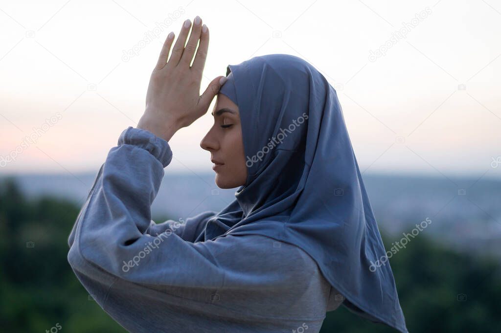 Young beautiful girl in a hijab prays and meditates on a background of sunset and landscape