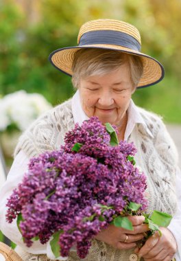 Şapkalı güzel, yaşlı bir büyükanne ellerinde bir demet leylak kokluyor ve bahçenin arka planında gülümsüyor.