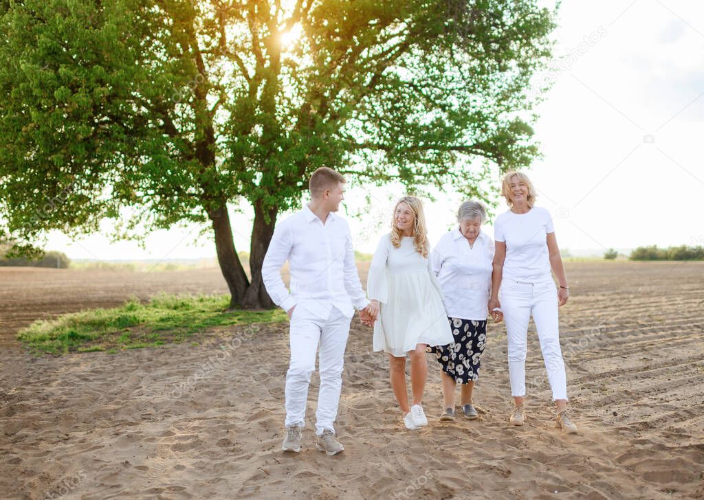 Family photo of mature mother, grandmother and eldest son and daughter in white clothes on the background of a single tree in a field at sunset