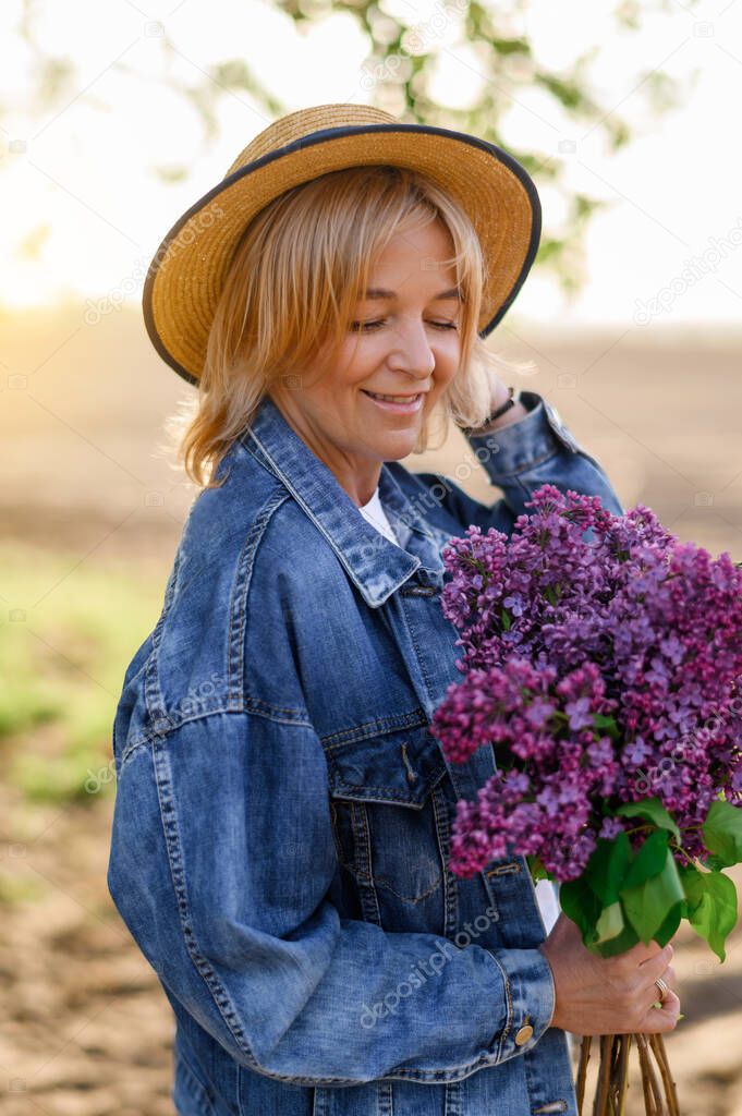 Portrait of mature beautiful caucasian blonde woman in hat and sniffing bouquet of lilacs at sunset on field background