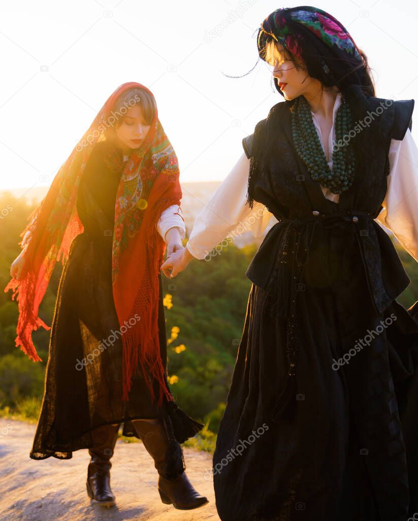 Beautiful young Caucasian girl in Ukrainian traditional national clothes holds her sisters hand and walks on the mountain at sunset