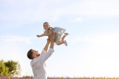 Yakışıklı beyaz bir baba ve küçük bir kız onu lup çiçekleriyle dolu bir tarlada günbatımının arka planına fırlatıyorlar.