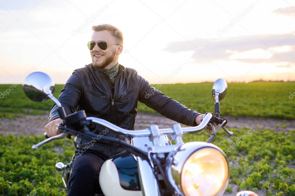 Young fair-haired brutal biker guy with a beard in sunglasses on a motorcycle in a field at sunset