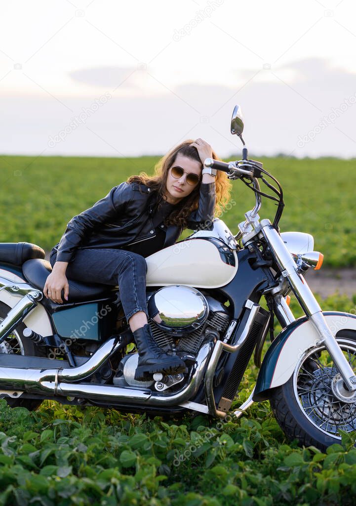 Young beautiful girl in leather clothes posing on a motorcycle in a field at sunset