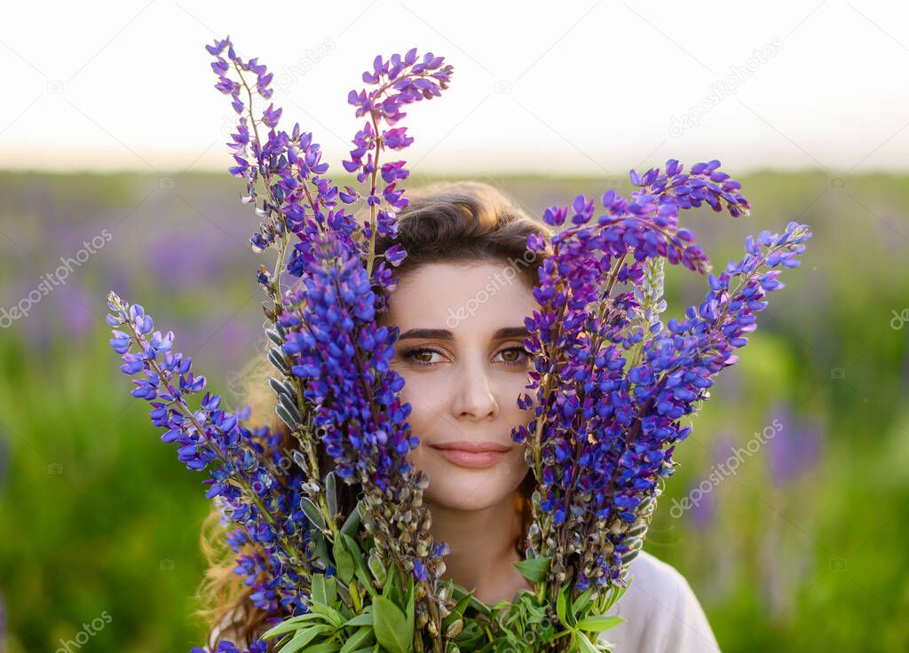 A young beautiful Caucasian girl poses with a bouquet of lupines against the background of the sunset in the field