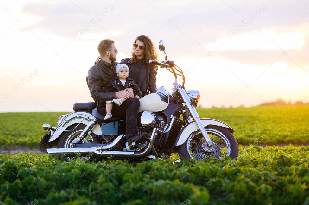 Young beautiful family dad mom and little daughter ride a motorcycle in the field at sunset and pose