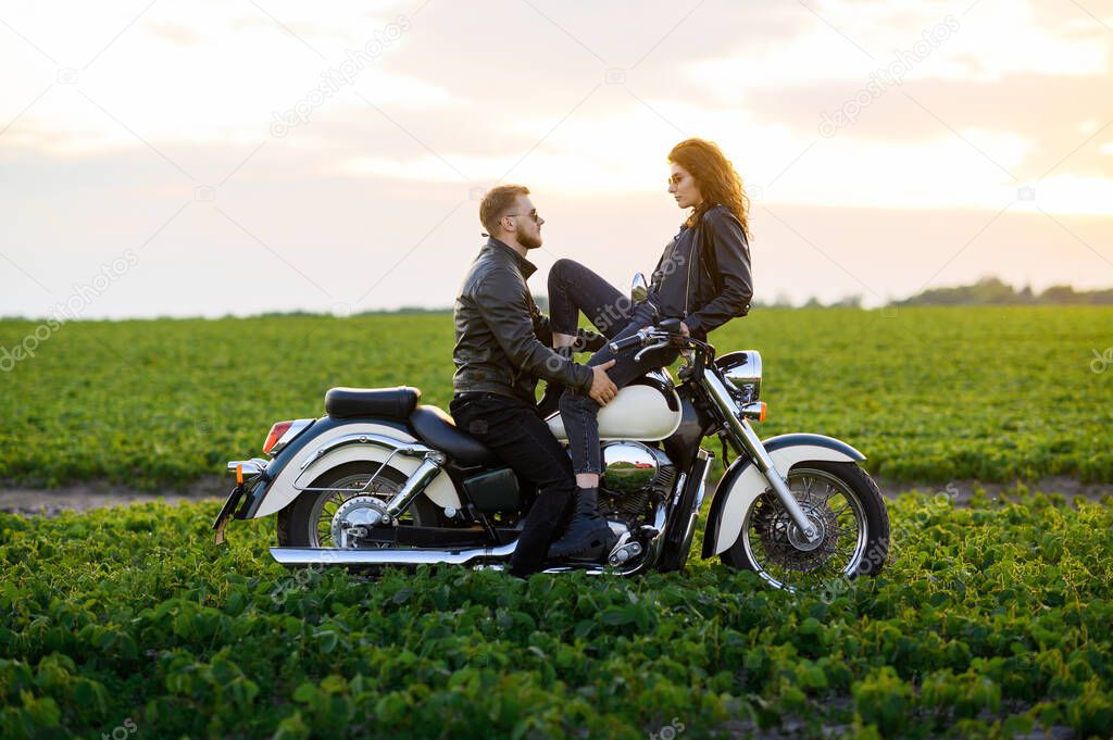 A young beautiful couple, a guy and a girl in leather clothes, are posing and hugging on a motorbike in a field against the background of the sunset