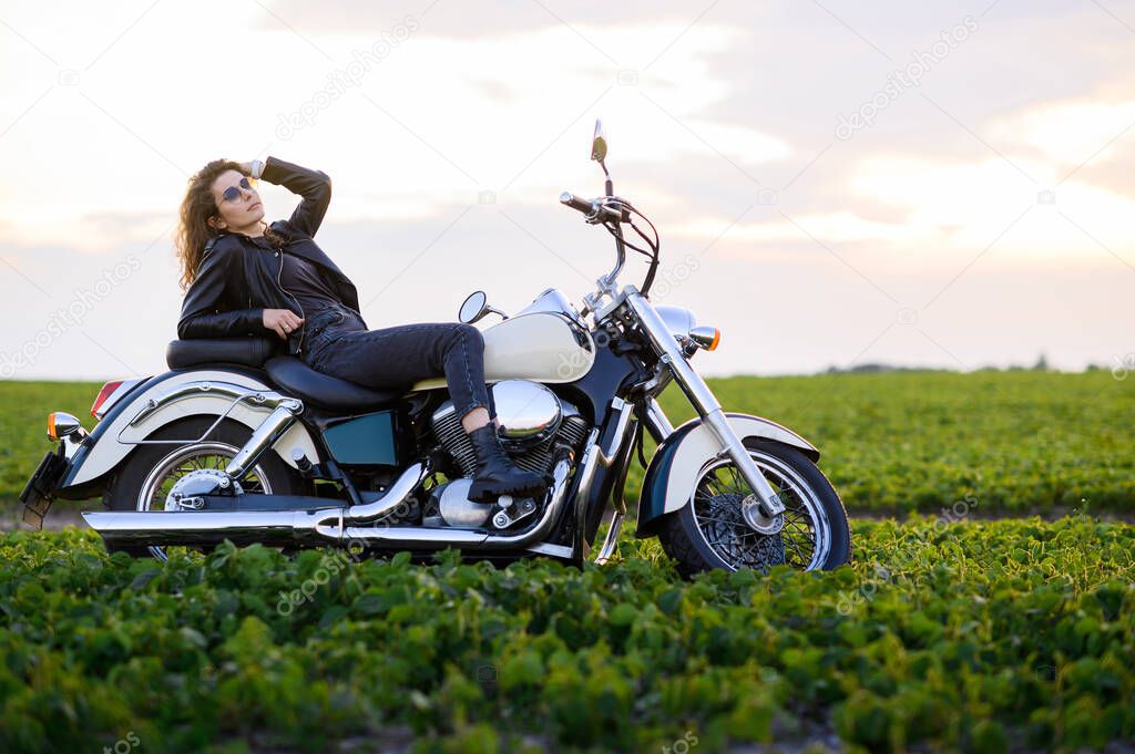Young beautiful girl in leather clothes posing on a motorcycle in a field at sunset