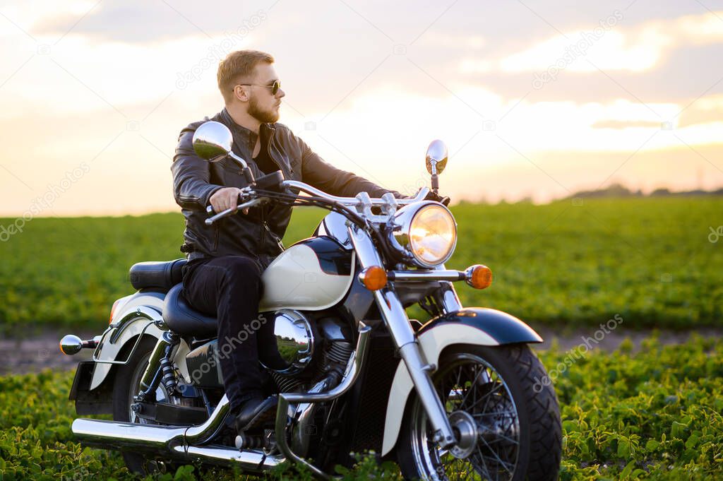 Young fair-haired brutal biker guy with a beard in sunglasses on a motorcycle in a field at sunset