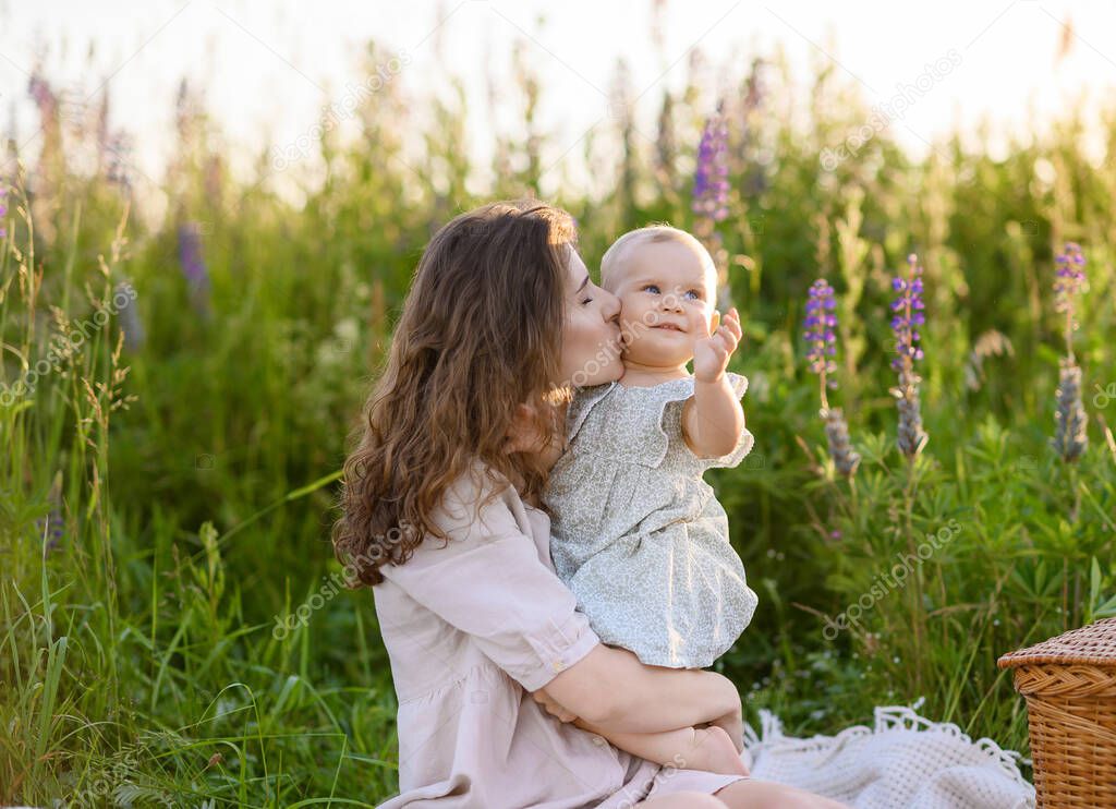 Young beautiful Caucasian mother is playing on a picnic with her little baby daughter against the background of the sunset in a field of lupines