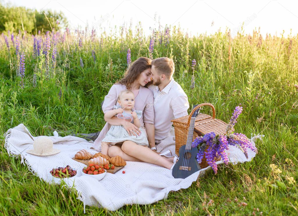 Beautiful Caucasian young family dad and mom with a little baby girl sitting on a picnic at sunset in a field of lupines